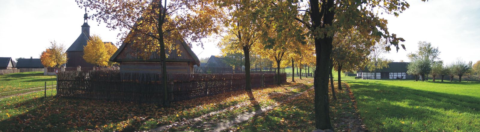 Fragment chałupy i Kościół z Wartkowic w tle w jesiennym pejzażu Wielkopolskiego Parku Etnograficznego w Dziekanowicach.